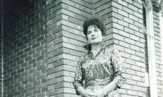 Singer, Lorri Gil dressed as Patsy Cline, standing at the corner of a brick building