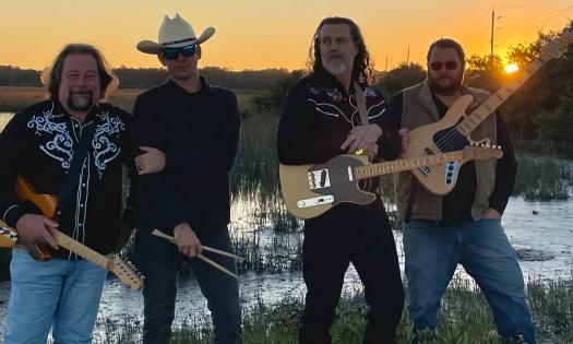The four members of the Oyster Bed Outlaws, standing with their instruments in front a local creek at sunset