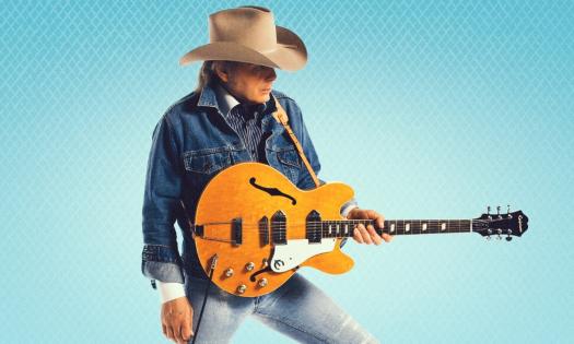 Dwight Yoakam strums his guitar and poses in denim jeans with a matching jacket. 