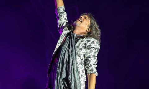 A Foreigner bandmember stands on stage and lifts one arm in the air. 