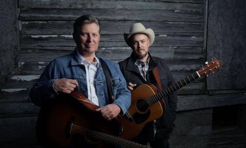 The Gibson Brothers pose with their guitars in front of a wooden background. 