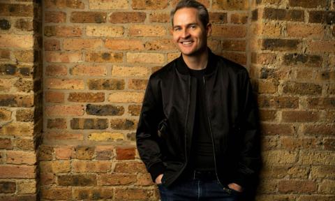 Pat McGann smiles and poses in a leather jacket in front of a brown brick wall. 