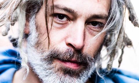 A headshot of Matisyahu wearing a blue shirt and posing in front of a white backdrop. 