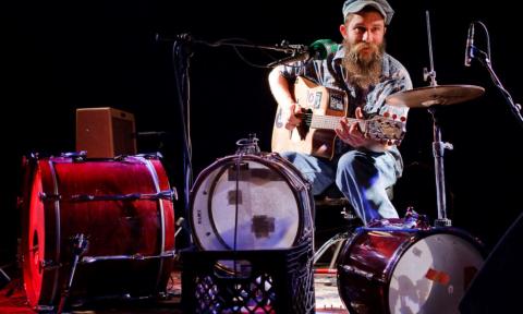 Ben Prestage wears a hat and holds his guitar as he sings on stage. 