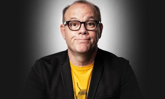 Tom Papa wears glasses and a black blazer and poses in front of a black and white backdrop. 