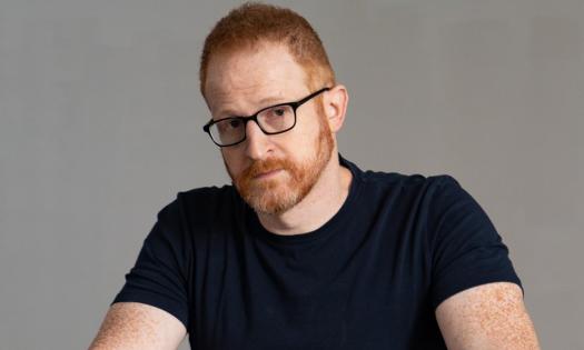 Steve Hofstetter wears glasses and a black t-shirt while posing in front of a grey backdrop. 