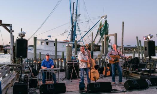 Bandmates from Blue Dogs sing and perform on a dock with a medium-sized boat and the open ocean as their backdrop. 