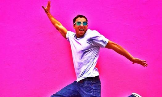 Brent Pella grins and jumps in front of a bright pink backdrop. 