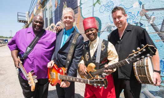 Lil Ed & The Blues Imperials smile and pose with their guitars to the side of a blue brick wall. 