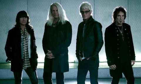 Bandmates from REO Speedwagon wear black clothing while posing in front of a well-lit backdrop. 