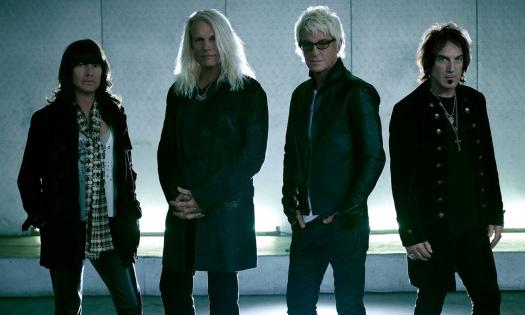 Bandmates from REO Speedwagon wear black clothing while posing in front of a well-lit backdrop. 