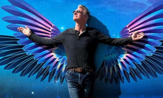Eliot Lewis smiles and poses in a black shirt and sunglasses while stretching his arms out in front of a blue backdrop with wings. 