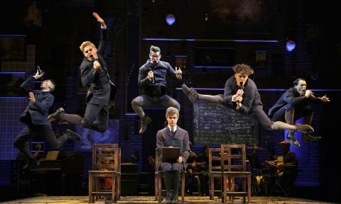 Six young men, in 19th Century costumes, leaping on the set of a school room