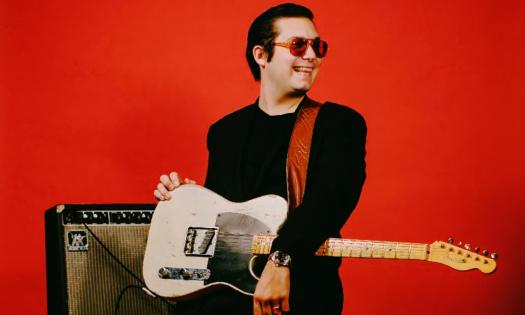 Eddie 9V holds his guitar and smiles in front of a red backdrop. 