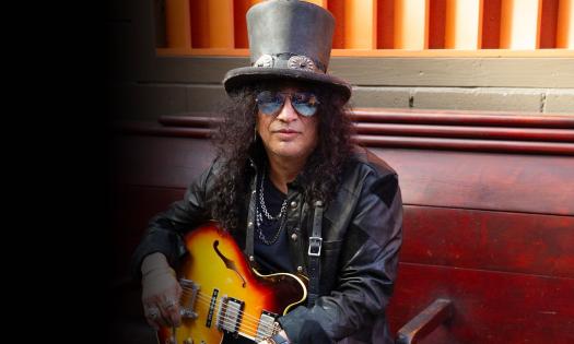 Slash poses with a black hat, matching leather jacket, and guitar. 