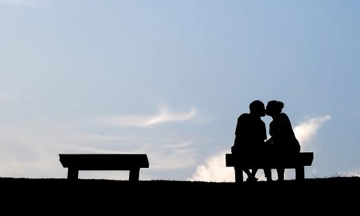 A couple kisses while sitting on a bench at the top of a hill in front of a lovely blue sky