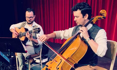 Jordan Taylor on guitar and Micah Taylor on cello, performing on stage in front of a red curtain