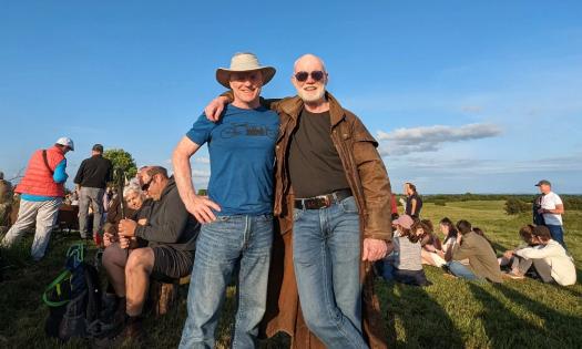 Two men stand on a hill, with several people ranged out behind them sitting and standing