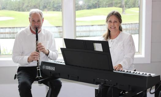 A musical duo, the man on clarinet, and the woman on keyboards, pose in front of a window