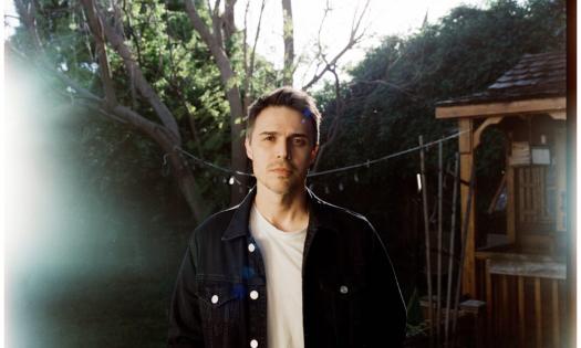 Kris Allen poses in a denim jacket in front of a tree and bushes. 
