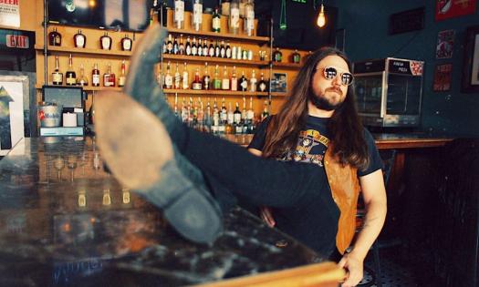 Sam Morrow poses with black glasses while sitting in front of a bar with wooden accents. 