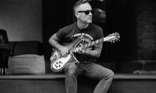 A black and white photo of Dave Hause strumming his guitar with a stage as his backdrop. 