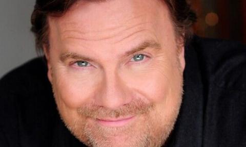 Kevin Farley smiles and poses in front of a grey background with bokeh. 