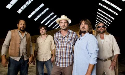 The members of Honey Island Swamp Band smile as they pose together inside a rustic barn. 