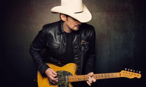 Brad Paisley wears a white cowboy hat and leather jacket while clutching his guitar in front of a black backdrop. 