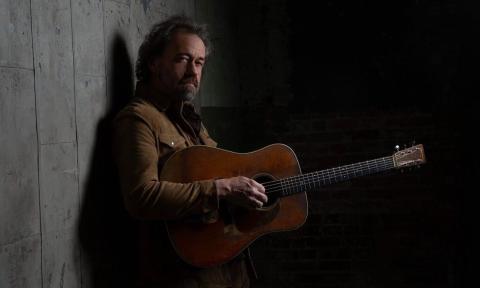 Dan Tyminski leans against a gray wall and strums his guitar.