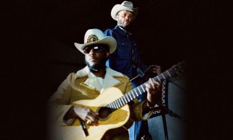 Leon Bridges strums his guitar while Charley Crockett poses in a white cowboy hat.