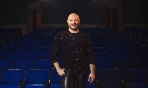 Travis Holp wears black clothing while posing in front of a row of blue chairs. 