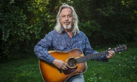 Jim Lauderdale poses with his guitar against a backdrop of lush green foliage.
