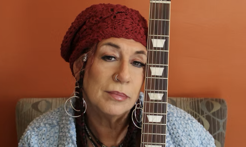 Joanna Connor wears a knitted hat and poses with her guitar in front of an orange backdrop.
