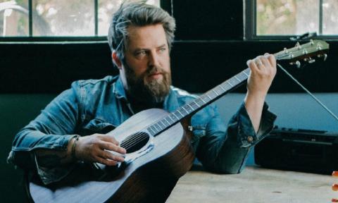 Lee Dewyze sits at a desk and strums his guitar.