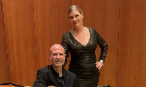A man, seated, and a woman, standing, against a wood-toned wall