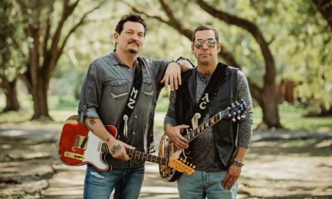 Mike Zito and Albert Castiglia stand together with their guitars in front of a tree-lined road.