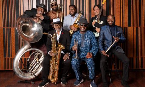 Members of the Dirty Dozen Brass Band smile and strike a pose in front of a rustic wooden backdrop. 