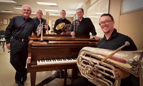 Five brass playing musicians pose around a piano with their instruments