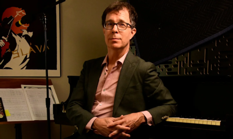 Ben Folds poses next to a piano and a microphone.