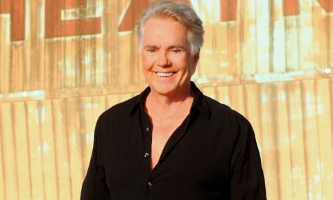 Shaun Cassidy wears a black shirt and poses in front of a gold backdrop.