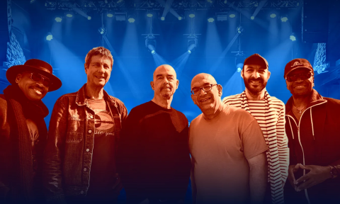 Bandmates from Acoustic Alchemy huddle together and pose with a stage as a backdrop.