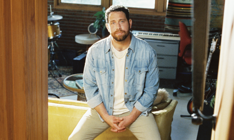 Jordan Fletcher strikes a relaxed pose on the edge of a couch, wearing a classic denim shirt.