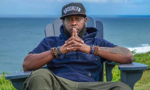Talib Kweli sits back in a chair musing in front of an open ocean. 