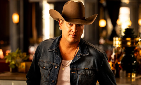 Jon Pardi poses with a brown cowboy hat and dark blue denim jacket in front of a bar with brown and amber hues. 