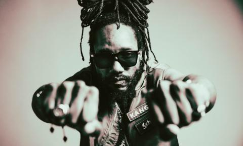 Kabaka Pyramid raises two of his fists and poses in front of a camera. 