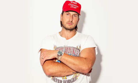 Russell Dickerson wears a bright red cap and folds his arms in front of a white backdrop.