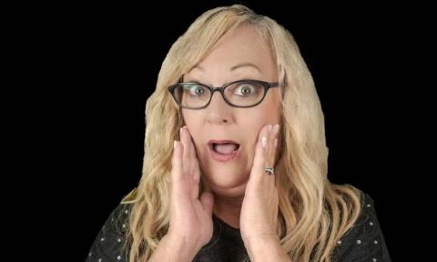 Shelly Belly poses before a black backdrop, hands pressed to her cheeks, her face lit with a look of wide-eyed surprise.