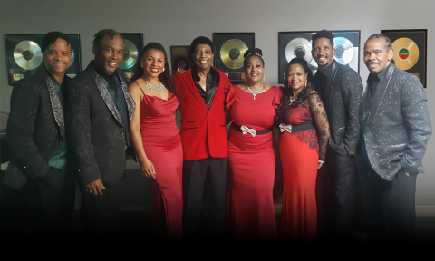 Bandmates from Forever Motown, dressed in black and red formal attire, pose proudly in front of a wall showcasing gold and platinum awards.