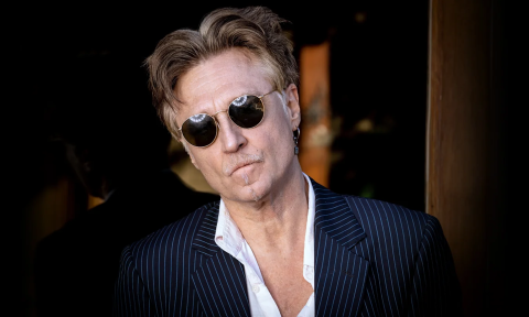 John Waite wears black glasses and poses in a pinstripe blazer with a white dress shirt.
